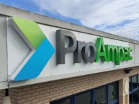 arge 3D fascia sign for ProAmpac, mounted to the exterior of a commercial building. The sign features substantial built-up letters with visible depth, projecting from a white fascia panel to create a bold three-dimensional effect. The "Pro" lettering is finished in dark grey, while "Ampac" is displayed in bright green, complemented by a raised geometric logo in green and blue. The combination of deep 3D lettering, strong corporate colours, and shadow effects creates a high-impact, contemporary branding display that stands out against the building façade and blue-framed windows below.