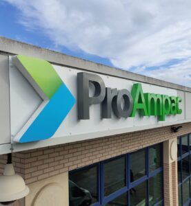 Large 3D fascia sign for ProAmpac, mounted to the exterior of a commercial building. The sign features substantial built-up letters with visible depth, projecting from a white fascia panel to create a bold three-dimensional effect. The "Pro" lettering is finished in dark grey, while "Ampac" is displayed in bright green, complemented by a raised geometric logo in green and blue. The combination of deep 3D lettering, strong corporate colours, and shadow effects creates a high-impact, contemporary branding display that stands out against the building façade and blue-framed windows below.