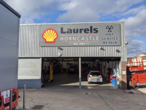 Large external fascia sign for Laurels Horncastle, mounted above the entrance to a vehicle service and repair centre. The sign features a dark grey background with prominent raised lettering spelling "Laurels Horncastle", accompanied by the distinctive Shell logo on the left. To the right, bold text highlights "MOT Service Repair" beneath a geometric automotive-style symbol. Red accent lines and subtle shadowing add depth to the design, while the oversized branding creates a professional and highly visible frontage that stands out against the corrugated metal building façade.