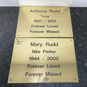 Pair of polished brass memorial plaques engraved with black text and mounted side by side on a dark stone surface. The plaques commemorate Anthony "Tony" Rudd (1941–2021) and Mary Rudd, née Parker (1944–2000), each featuring the inscription "Forever Loved, Forever Missed". Small interlocking heart symbols are engraved in the corners, adding a decorative touch. The reflective brass finish contrasts with the dark engraved lettering, creating an elegant and dignified memorial display.