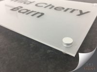 A close up of a modern, frosted acrylic sign for Wild Cherry Barn, mounted on brushed aluminium stand-off fixings. A circular aluminium stand-off cap secures the panel at the corner, creating a subtle floating effect that gives the signage a clean, contemporary appearance.