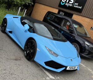 Bright blue Lamborghini Huracán parked outside the GRS Signs premises. The sports car features a sleek blue bodywork finish with black alloy wheels and a black roof. In the background, a GRS Signs-branded commercial van and the company's external fascia sign are visible, showcasing the business premises.