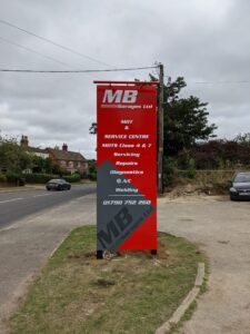 Freestanding modular sign for MB Garages Ltd, featuring a bold red panel mounted within a black sign frame. The sign displays the MB Garages Ltd logo in white at the top, followed by a list of services including MOT, Service Centre, MOTs Class 4 & 7, Servicing, Repairs, Diagnostics, A/C, and Welding. White italic typography and angular grey graphic accents add a dynamic, automotive-inspired appearance, while the contact telephone number is displayed prominently at the bottom. The vibrant red colour scheme and tall vertical format create a highly visible roadside advertising sign.