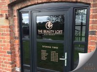 The Beauty Loft Retreat, featuring elegant gold vinyl graphics applied to the front door's glazing. The branding includes a stylised leaf logo above the business name “THE BEAUTY LOFT RETREAT” in large capital letters. Below, a section titled “OPENING TIMES” lists the business hours. The dark grey door frame and silver door handle contrast with the gold lettering - with the overall design looking clean, sophisticated, and well suited to a professional beauty and wellness business.