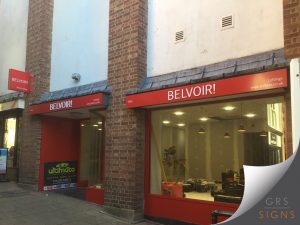 Exterior storefront signage for Belvoir!, featuring bold white lettering on a bright red fascia mounted above large shopfront windows. The signage spans multiple sections of the brick-fronted premises, with a projecting red wall-sign also at at the corner of the photograph, mounted to the building.