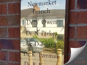 Brass name plaques located on an external wall and fixed to a wooden frame, listing the various medical practitioners that work at Newmarket Branch (Louth) in engraved black lettering.