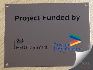 An engraved bronze plaque with circular, drilled fixing holes. Engraved black letters read "Project Funded by" - and the HM Government logo and "Greater Lincolnshire Local Enterprise Partnership" logos sit towards the bottom.