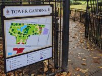 Freestanding park information and wayfinding sign for Tower Gardens, mounted to decorative black metal railings. The sign features a white header panel with the words "TOWER GARDENS" in bold black lettering, alongside council branding. Below, a colour-coded site map illustrates the layout of the gardens, including pathways, green spaces, water features, entrances, and key visitor facilities. A legend at the bottom provides symbols and directional information to help visitors navigate the park. The sign combines a cream, green, blue, and red colour palette with clear graphics and typography, creating an attractive and easy-to-use visitor information display.