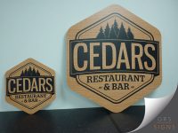 Two bespoke wooden signs for Cedars Restaurant & Bar displayed against a light blue background. The signs are hexagonal with rounded corners and feature a natural wood-effect finish engraved with dark contrasting graphics and text. Both signs display the CEDARS logo in large bold lettering, with a silhouette of evergreen trees above and the text “Restaurant & Bar” below. The larger sign is positioned to the right, with a smaller matching version to the left, demonstrating the design at different sizes. The rustic wood finish and engraved detailing create a warm, premium appearance.