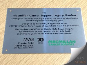 Brushed stainless steel commemorative plaque with chemical etching, for the Macmillan Cancer Support Legacy Garden at Chesterfield Royal Hospital NHS Foundation Trust. The plaque features engraved black text explaining the garden's purpose as a space for reflection and recognising the importance of legacy gifts. It notes that the garden was designed by Lara Behr, won a gold medal at the 2017 RHS Tatton Park Flower Show, and was gifted to the hospital by Macmillan Cancer Support. Logos for NHS Chesterfield Royal Hospital, 70 Years of the NHS, and Macmillan Cancer Support are displayed along the bottom. The plaque is able to be mounted with metal fixings via the circular drill holes.