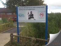 A blue dual-post mounted external sign for the NHS, encouraging no smoking on site at this hospital in Chesterfield. On the left is a blue “Proud to be Smoke Free” logo, while the centre includes a black-and-white photograph of a healthcare worker. The white sign is mounted between two blue posts and features the headline "Don’t be offended by our wish to keep people healthy and our hospital clean." On the right is a no-smoking symbol alongside text explaining that the hospital operates a smoke-free site. Additional information at the bottom promotes smoking cessation support through Live Life Better Derbyshire, including a freephone number and website details. The sign is positioned outdoors, beside a hospital car park.