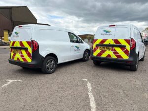 Two white East Lindsey District Council vans with printed vehicle graphics, including the East Lindsey District Council's logo and reflective red and yellow rear chevrons. The vans are parked outside.