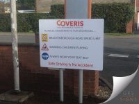 White dual-post mounted roadside sign for Coveris High Performance Packaging. The sign displays a series of road safety messages, including reminders about the 30 mph speed limit, children playing, and always wearing a seatbelt, alongside relevant safety symbols. Red text at the bottom reads "Safe Driving Is No Accident". The sign is positioned at a site entrance beside a brick wall and public road. Text is mainly written in black or bold, red and underlined.