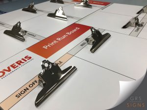 Close-up of a large print run board used for packaging proofing and sign-off. Several shiny metal bulldog clips are attached across a white grid layout containing labelled sections for samples, reel information, and approvals. A prominent orange-red panel in the centre reads “Print Run Board” in white text. The lower-left corner displays the NOVERIS Performance Packaging logo, while a beige strip labelled “SIGN OFF” runs across the board.