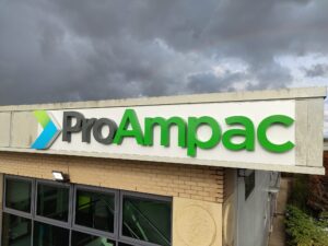 Large 3D fascia sign for ProAmpac, mounted to the exterior of a commercial building. The sign features substantial built-up letters with visible depth, projecting from a white fascia panel to create a bold three-dimensional effect. The "Pro" lettering is finished in dark grey, while "Ampac" is displayed in bright green, complemented by a raised geometric logo in green and blue. The combination of deep 3D lettering, strong corporate colours, and shadow effects creates a high-impact, contemporary branding display that stands out against the building façade and blue-framed windows below.
