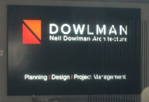 Illuminated 3D wall sign for Neil Dowlman Architecture, featuring a sleek black fascia with the company's distinctive orange and red geometric logo. White illuminated lettering displays "DOWLMAN" and "Neil Dowlman Architecture", creating a striking contrast against the dark background. Beneath, illuminated text highlights the firm's core services: "Planning | Design | Project Management".