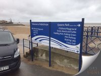 Large blue wayfinding sign on a seaside promenade in Mablethorpe, mounted on three blue posts beside a concrete sea wall. The sign welcomes visitors to Queens Park and provides directional information to local attractions, including the Children's Playground, Tennis Courts, Bowls, Cafés, Paddling Pool, Putting Green, Mini Railway, Boating Lake, and Crazy Golf. White arrows indicate directions to various destinations, including North Promenade and Central Promenade. A stylised white wave graphic runs across the lower section of the sign, and the East Lindsey logo appears in the bottom-right corner. Sandy beach, sea-defence railings, and the coastline are visible in the background.