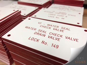 Stacks of engraved, industrial valve identification tags - white background with a red core. The tags feature precision-engraved red text to aid with identifying equipment and lock-off points. The signs visible in the image include details such as "V2 - Water Seal Check Valve", "Water Seal Check Valve Drain Valve", and "Lock No. 149". Each tag includes pre-drilled fixing holes for installation.