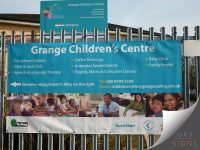 A large banner, attached via reinforced eyelets to outdoor fence railings, to advertise Grange Children's Centre. It features a turquoise background, white text, and photographs of smiling adults & children in a learning environment.