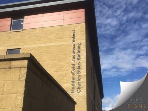 The exterior of a building showing cut-out, large lettering in a brushed metallic material and 3D-style, to identify Huddersfield Business School Charles Sike Building. The text is displayed on its side and stands out against the light sandstone coloured bricks.