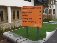 Dual-post, double-sided wayfinding sign at University of Huddersfield, showing where graduates need to be during graduation ceremonies. The sign points towards registration, robing, photography and drinks reception. The sign has an orange background - featuring bold, black lettering - and the University's typography logo displayed at the bottom right in white.