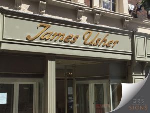 Positioned above a shop entrance: 3D cut-out gold, elegant lettering in a scripted/cursive font to identify James Usher jewellers. The gold lettering looks sleek standing out from the sage green paint behind.