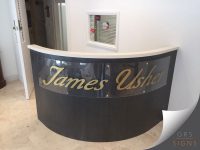 Bright, modern jewellery showroom interior featuring bespoke branded signage for James Usher. A curved reception desk forms the focal point of the space, displaying the name “James Usher” in elegant gold script lettering on curved Perspex across the front.