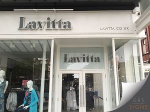 Modern retail unit for Lavitta during a store fit-out, featuring a clean, contemporary shopfront with a large "Lavitta" logo displayed on a light grey fascia (which can be illuminated). Large floor-to-ceiling display windows on either side of the entrance contain promotional posters announcing "NEW STORE COMING SOON!" and describing the business as "Fashion For Real Women." Bright interior lighting and a minimalist colour scheme create a modern retail appearance.