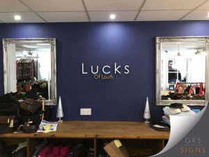 A navy blue interior wall with 3D cut out lettering for "Lucks Of Louth". "Lucks" is larger and the letters spread out slightly, coloured in white, with "Of Louth" underneath, smaller and coloured with a golden yellow shade. Two large, ornate silver mirrors are positioned either side.