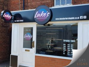 Exterior shopfront signage for Luke's Barbershop, featuring a sleek dark navy fascia with circular projecting signs displaying the business logo in retro-inspired pink, blue, and white colours. The main fascia includes the company website, social media icons, and contact number in white lettering, while matching branding is applied to the entrance door and window. White opening hours graphics are displayed on the front window, creating a professional and cohesive storefront design.