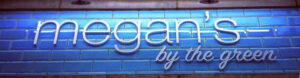 An illuminated storefront sign for "megan's by the green". The cut out, 3D white lettering stands out from the tiled blue background. "megan's" is in a contemporary font, with "by the green" being in a smaller, flowing script style font. The lighting installed illuminates the white lettering - showing a nice contrast against the rich blue tiles.