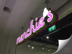 Illuminated projecting shop sign suspended from the ceiling. The three-dimensional sign features bright pink lowercase lettering outlined in white, with internally illuminated faces creating a vibrant glow. The sign is mounted on a deep green backing panel with a contrasting pink edge, giving it a bold, layered appearance. A green emergency exit sign is visible behind the display, while the surrounding ceiling and retail unit interior are lit by overhead spotlights. The eye-catching design creates strong visibility and reinforces the brand's playful, modern identity.