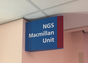 A modular projecting sign mounted to an internal wall, used to identify the NGS Macmillan Unit. The sign is NHS blue with white bold lettering and a red edge.