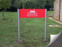 A dual-post, mounted entrance sign for Oadby Food Court. The sign features a vibrant red rectangular panel with the Oadby Food Court logo and name displayed in white lettering. A stylised building graphic appears above the text, creating a modern and professional visual identity. Trees and open green space are visible in the background.