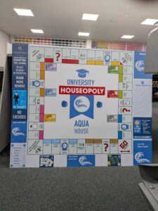 Bespoke educational wall display designed as a custom "University Houseopoly" board game. The large modular sign features colourful game-board-style panels, house points information, school values, rewards and behaviour guidance, creating an engaging and interactive display for a school environment.