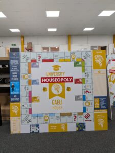 Bespoke educational wall display designed as a custom "University Houseopoly" board game. The large modular sign features colourful game-board-style panels, house points information, school values, rewards and behaviour guidance, creating an engaging and interactive display for a school environment.
