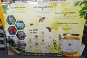 A brightly coloured modular educational display board about bees, gardening and sustainability stands in a workshop environment. The sign features hexagonal photo panels showing flowers and raised garden beds, alongside illustrations of bees and a honey jar. Headings include "From our Garden", "Pollinated with Care", "From Hive to Home" and "Sustainable at Home", connected by dotted flight paths. An information section titled "The Buzz on Bees" highlights facts about pollination and honey production. The design uses soft green and yellow tones with floral graphics and promotes environmental awareness and school-grown produce.