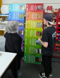 A large, colourful timeline display board is being positioned in a workshop by a person wearing a black GRS Signs branded T-shirt and cap. The vertical panel features historical periods with colour-coded sections, images and text describing key eras.