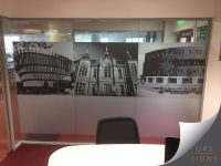 Large glass partition graphic, printed/etched and frosted, installed within an office building. It features a three black-and-white photographic panels applied across the glazing. The images showcase a mix of modern and historic architecture, including a contemporary curved building, an ornate Victorian-style building with decorative stonework and pointed roofs, and a modern multi-storey building with landscaped surroundings. The graphics are positioned in a horizontal strip across the centre of the glass partition, with frosted lower panels providing additional privacy. Office furniture, doorways, and interior spaces are visible beyond the glass, creating a professional and contemporary environment. The monochrome imagery adds visual interest to the space.
