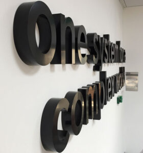 Bold font, lower-case 3D black lettering - displayed internally as wall art and reads "one system for a complex world". The letters have depth and project from the wall, creating a striking dimensional effect - emphasised by the angle of the photograph.