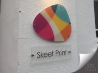 A close-up of 'Skeet Print' wall signage logo, which features a vibrant, irregularly shaped acrylic logo panel mounted on stand-off fixings, secured to a white rendered wall. The logo combines overlapping colours including pink, orange, yellow, teal, blue, and cream. Beneath the logo, a clear plaque-style sign is mounted with metal stand-off fixings and displays the company name "Skeet Print" in simple black lettering.
