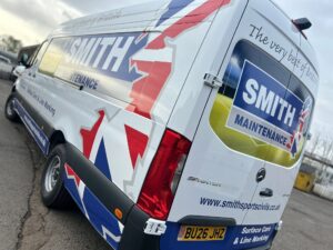 Rear three-quarter view of a white Mercedes Sprinter van featuring a striking Smith Maintenance vehicle livery. The design incorporates large blue and white company branding, red, white and blue Union Jack-inspired graphics across the side and rear panels, and printed contact information including website details.