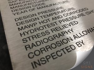 An angled close up of a brushed, stainless steel nameplate with black lettering. The plaque includes fields such as Manufacturer, Design Code, Year of Manufacture, Design Pressure, Design Temperature, MAWP Hot and Cold, Hydrotest Pressure, Stress Relieved, Radiography, Corrosion Allowance, and Inspected By.