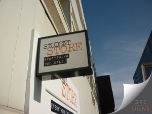 A wall-mounted, double-sided sign, projecting from the exterior of a building to ensure visibility from both sides. The sign features a white face with distressed-style lettering reading "STUDENT STORE", with "Student" in black and "Store" in orange. Beneath, the slogan "EVERYTHING YOU NEED" is displayed in white text on black rectangular panels. The sign is housed within a black metal frame - perfect for wayfinding and clearly identifying the store's location.