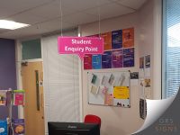 Suspended rectangular wayfinding sign reading "Student Enquiry Point" in bold white lettering on a bright pink background. The sign has rounded corners and a downward-pointing speech-bubble style marker beneath the text, indicating the location of the enquiry desk below. It is suspended from the ceiling by two thin metal cables within an indoor office or reception area.