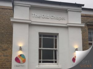 Exterior building signage for The Old Chapel, featuring raised white individual letters mounted directly onto a cream-coloured exterior wall. Below, a colourful circular logo sign for Skeet Print is displayed beside a modern wall-mounted up-and-down light fitting.