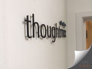 Light/narrow font display of 3D black lettering - displayed internally as wall art and reads "thoughtmix". The letters project from the wall, creating a striking dimensional effect - emphasised by the angle of the photograph. To the right of the text, three small circular graphic elements form part of the company logo.