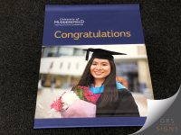 Promotional University of Huddersfield graduation flyer, featuring the university logo and the tagline "Inspiring tomorrow's professionals" on a dark blue background. The banner displays the word "Congratulations" in large gold lettering above a photograph of a female graduate wearing academic robes and a mortarboard, holding a bouquet of flowers.
