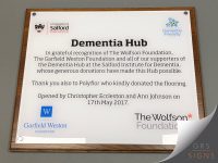 Wall-mounted donor recognition plaque for the Dementia Hub at the University of Salford Manchester. The plaque features black text on a white background, thanking the Wolfson Foundation, Garfield Weston Foundation, and other supporters for their contributions to the dementia facility. It also acknowledges a flooring donation from Polyflor and notes that the hub was opened by Christopher Eccleston and Ann Johnson on 17th May 2017. The plaque displays the University of Salford Manchester logo, the Dementia Friendly logo, and the logos of the Wolfson Foundation and Garfield Weston Foundation. Mounted with metal stand-off fixings, the plaque is presented within a wood-effect surround.