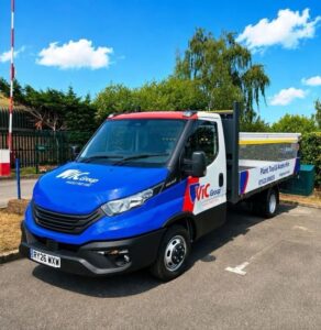 VIC Group branded flatbed hire truck parked outdoors on a sunny day, viewed from the front side. The vehicle features plant, tool and access hire branding, contact details and a mesh-sided load bed, with trees and a blue sky in the background.